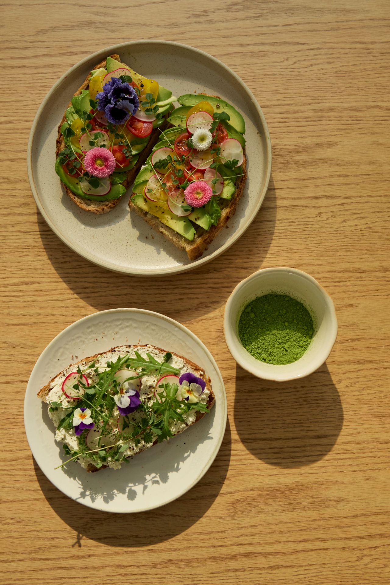 Two seeded toasts with avocado, radish and edible flowers beside a bowl of matcha on oak